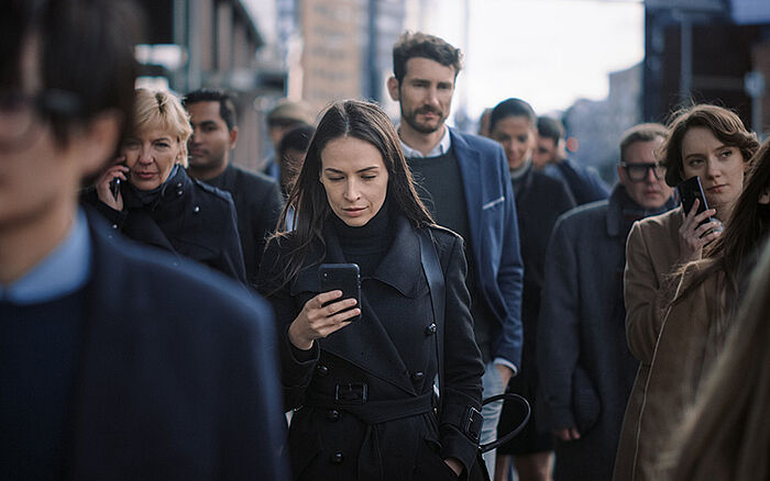 A stylish businesswoman in a black coat uses her smartphone while walking down a busy downtown pedestrian street, looking confident and engaged with her device.