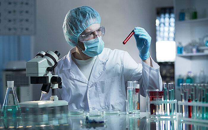 Laboratory worker analyzing blood samples in test tubes, wearing protective gear and using medical equipment, representing medical research or diagnostic testing.
