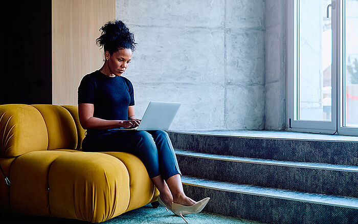 Focused professional using a laptop on a yellow sofa in a contemporary office setting.