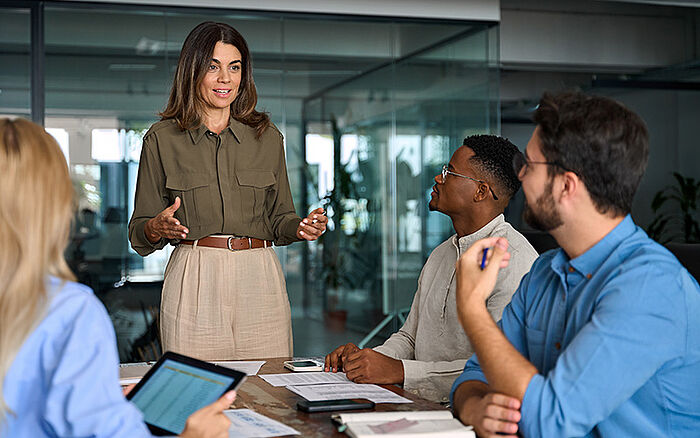 Business team meeting in a boardroom, with a woman leading a discussion on project planning and strategy alongside colleagues in a corporate office.