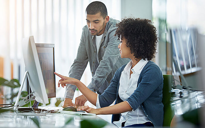 Two colleagues collaborating at a computer in a modern office setting. A man gestures toward the screen while a woman listens attentively, suggesting teamwork, guidance and digital problem-solving.