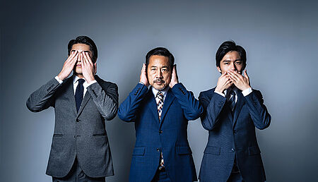 Three businessmen in suits, each covering either their eyes, ears or mouth with their hands, symbolizing the concept of “see no evil, hear no evil, speak no evil” in a corporate setting.