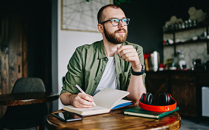 Bearded man sitting in a stylish café, writing ideas in a notebook.