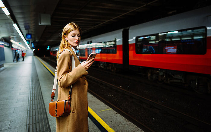 Young woman standing on a metro station platform looks at her phone in concern.