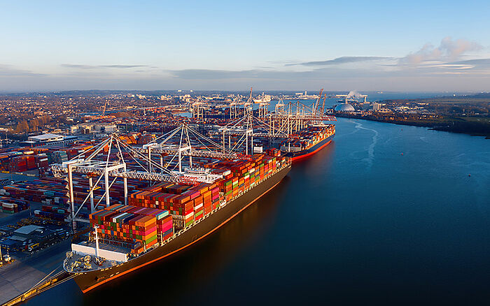 Aerial view of cargo ships docked at the Port of Southampton, with colorful shipping containers stacked on deck and along the terminal, showing cranes and port infrastructure.