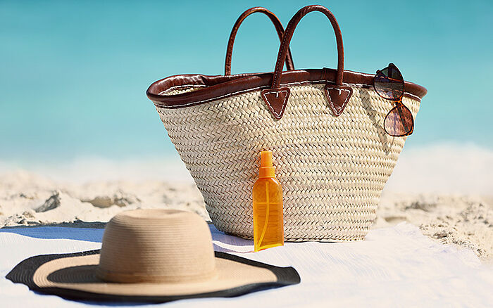 Women’s beach accessories including a straw tote bag, sun hat, and sunscreen bottle on sand with a blue ocean in the background.