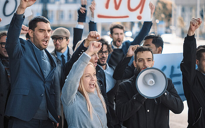 A diverse group of business people and office managers picketing on a street during a strike, holding signs and posters and one person using a megaphone, expressing demands for justice amid an economic crisis.