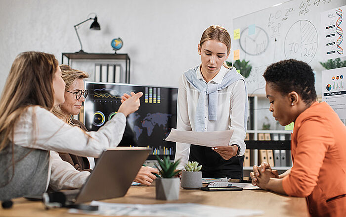 Young businesswoman in a white shirt smiling while presenting a financial report during a conference. She is standing at the head of a table with multiracial colleagues seated around her, reviewing documents and using laptops in a modern office setting.