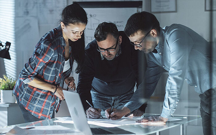 Three design engineers in a meeting reviewing technical drafts, components and a notebook, suggesting collaboration on confidential documents or trade secrets related to product development.