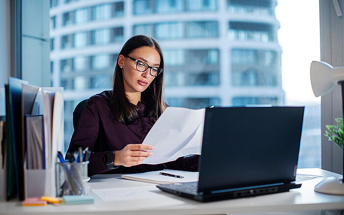 Focused woman reviewing financial documents at her desk in a modern office, surrounded by paperwork, a laptop and a calculator, suggesting accounting or administrative work.