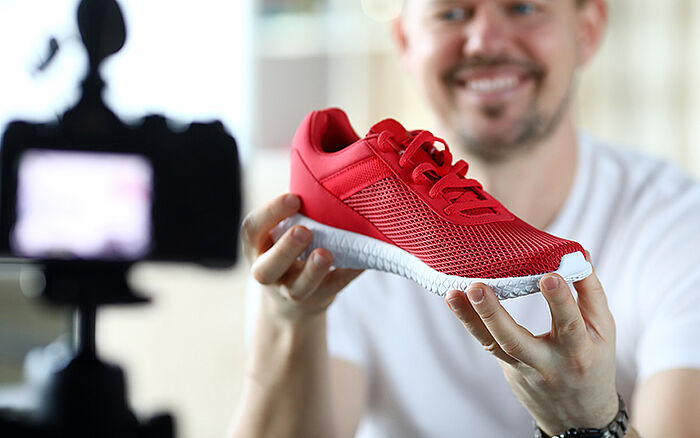 Man recording an online product review, holding a sports sneaker close to the camera while explaining how to spot authentic and fake pairs.